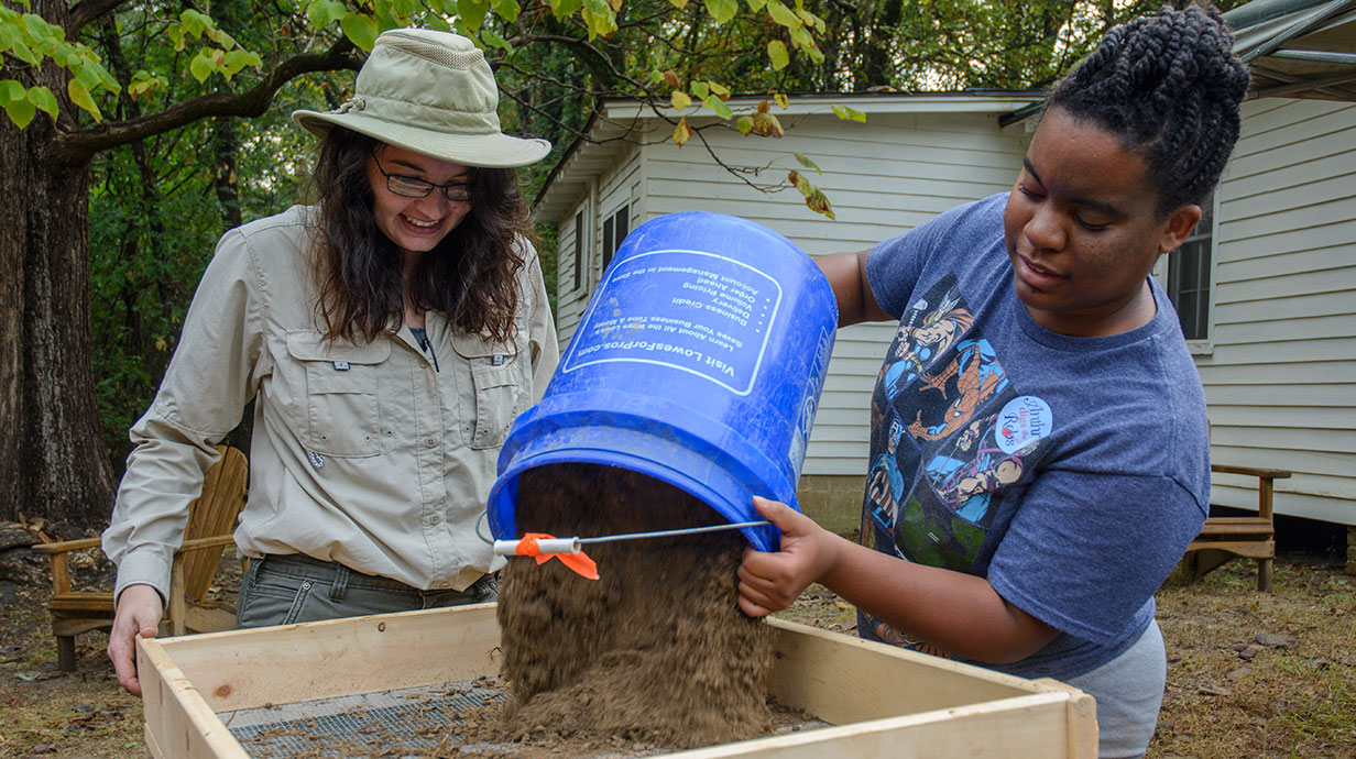 two women dupping dirt from a bucket into a net outdoors