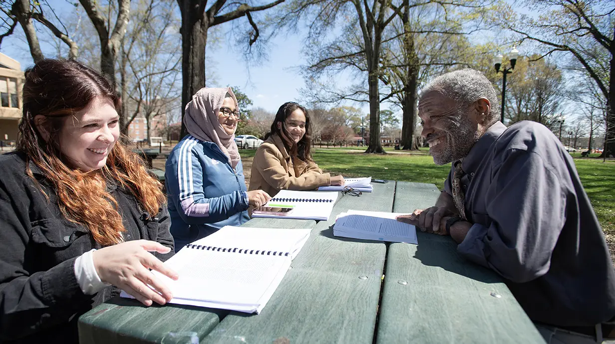 Three students and a professor sitting at a green picnic table outside. Everyone has an opened notebook or book in front of them.