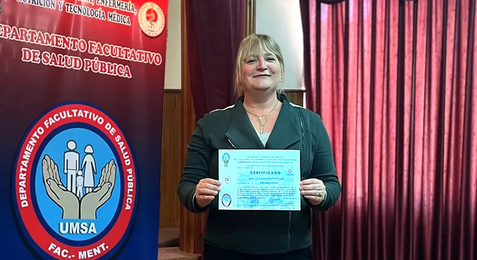 person smiling stands with certificate in hand. Banner to the left of person has text in Spanish.