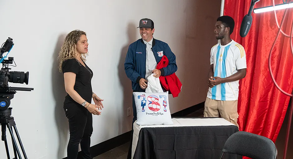 Three people standing in conversation around a small table and are flanked by a camera on a tripod and microphone