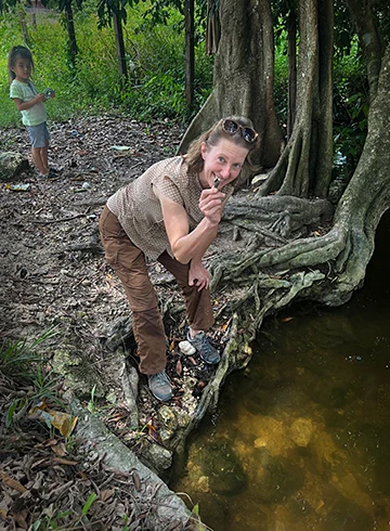 Professor leaning over looking at the camera holding a shell near a forested river bank. Child stands in background