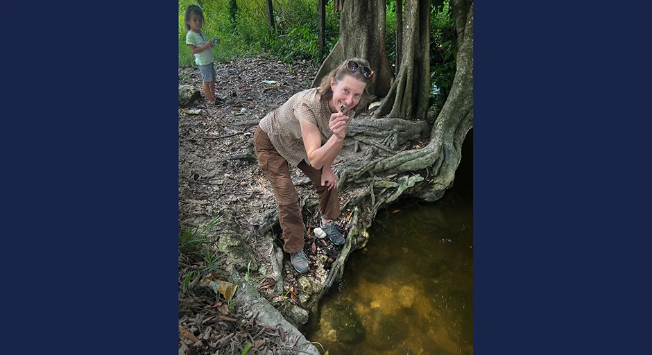 Professor leaning over looking at the camera holding a shell near a forested river bank. Child stands in background