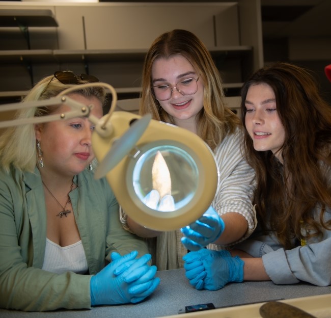 three students gathered together and looking at an arrowhead with a ring light and magnifying glass