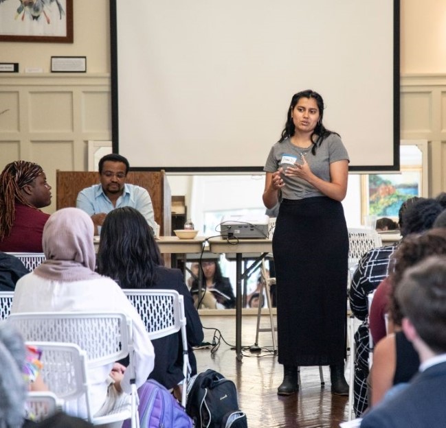 a woman standing in front of a seated group of people while making a presentation