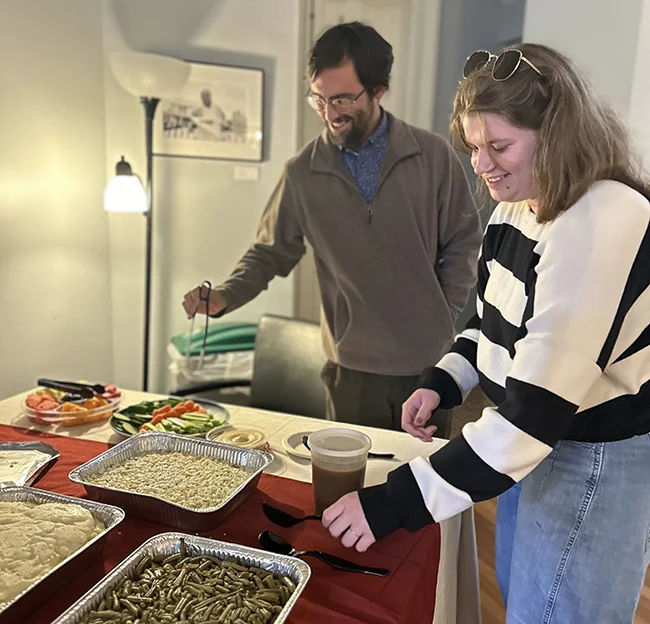 Two people stand over trays of food serving themselves