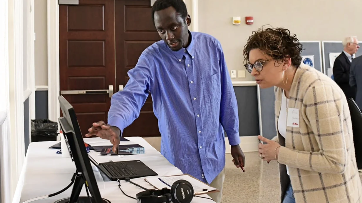 person standing looking at computer screen while another person gestures towards the screen. Headphones are attached to computer