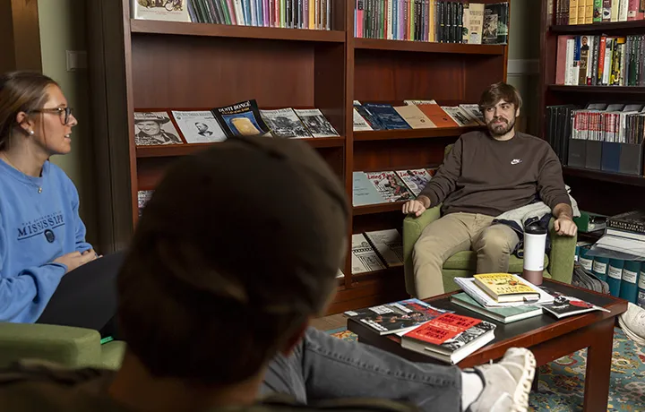 3 students sitting in lounge with background of books on bookshelves. 