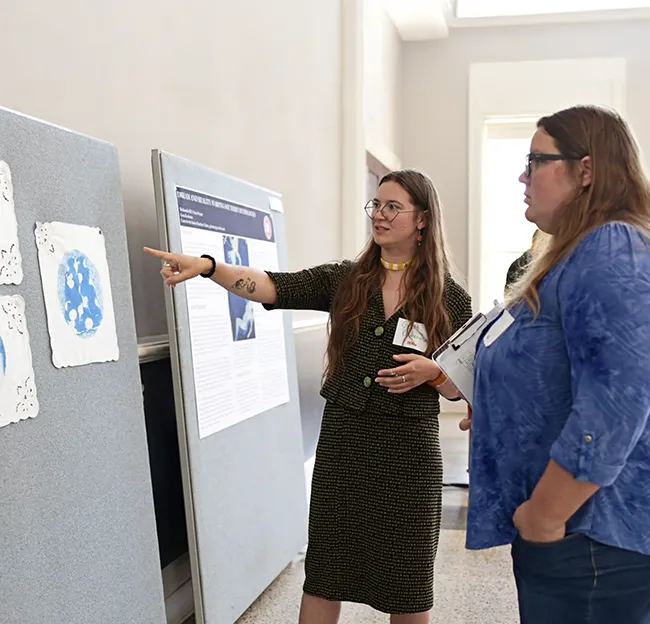 two people sitting standing in front of a display; one person points to display