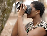Person in profile holding camera with big lens upward. Tree in background