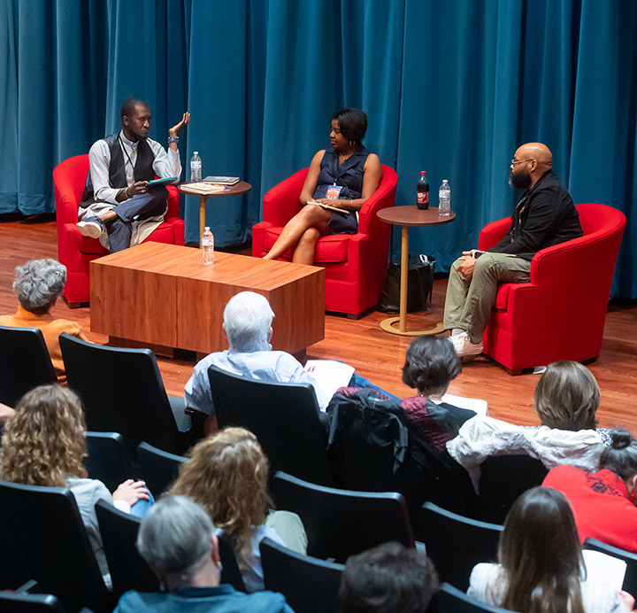 3 people sitting in red chairs speaking to a crowd on stage