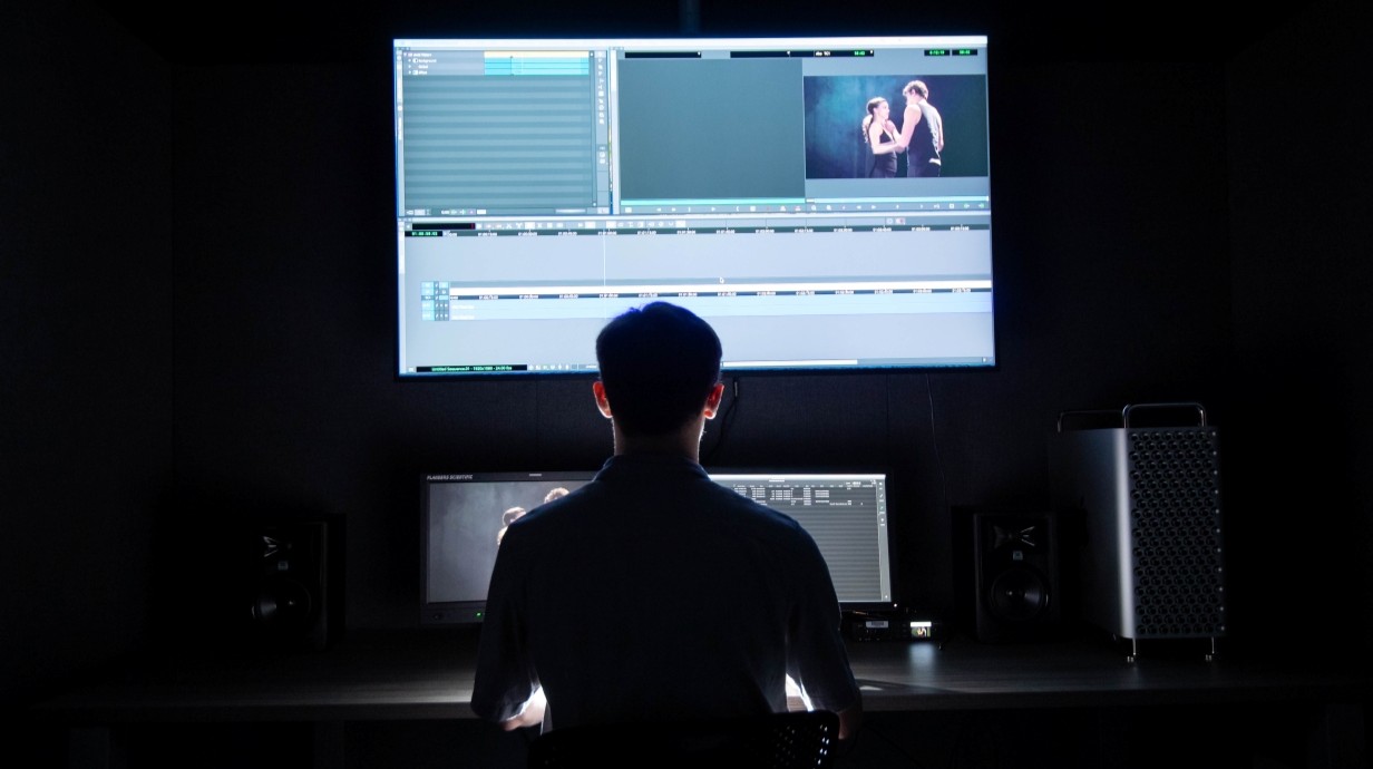 student seated in a dark room at a computer editing work station with a film showing on the big TV