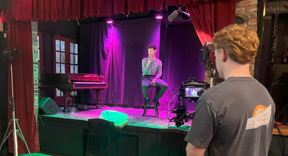 student seated on a stage holding a microphone while being filmed