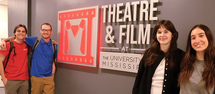 four students standing in front of a big sign on the wall that says "UM Department of Theatre & Film"