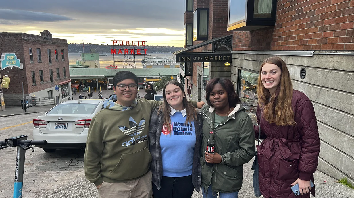 four college students smiling and standing in the street with Seattle's Public Market sign in background