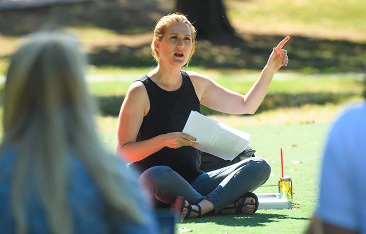 woman sitting outside cross-legged and pointing, students out of focus in foreground