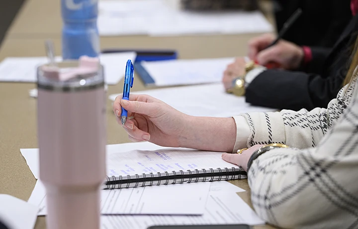 close up of hand holding pen hovering over paper. Pink cup on left