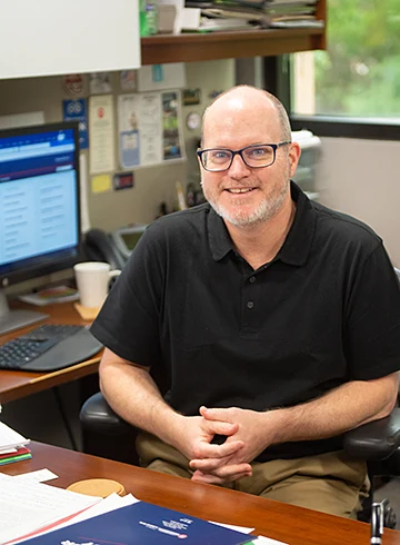 stephen monroe sitting at desk; computer in background and papers on desk