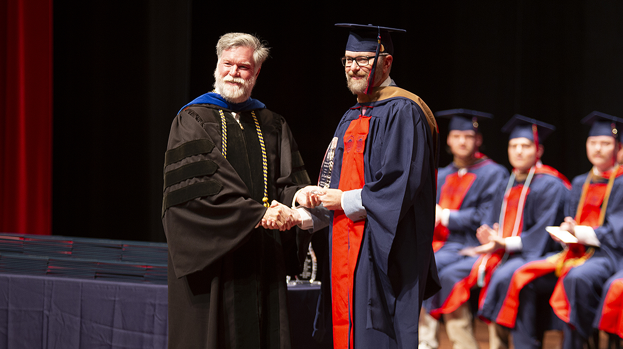 A man dressed in academic regalia hands a diploma to another man wearing graduation robes.