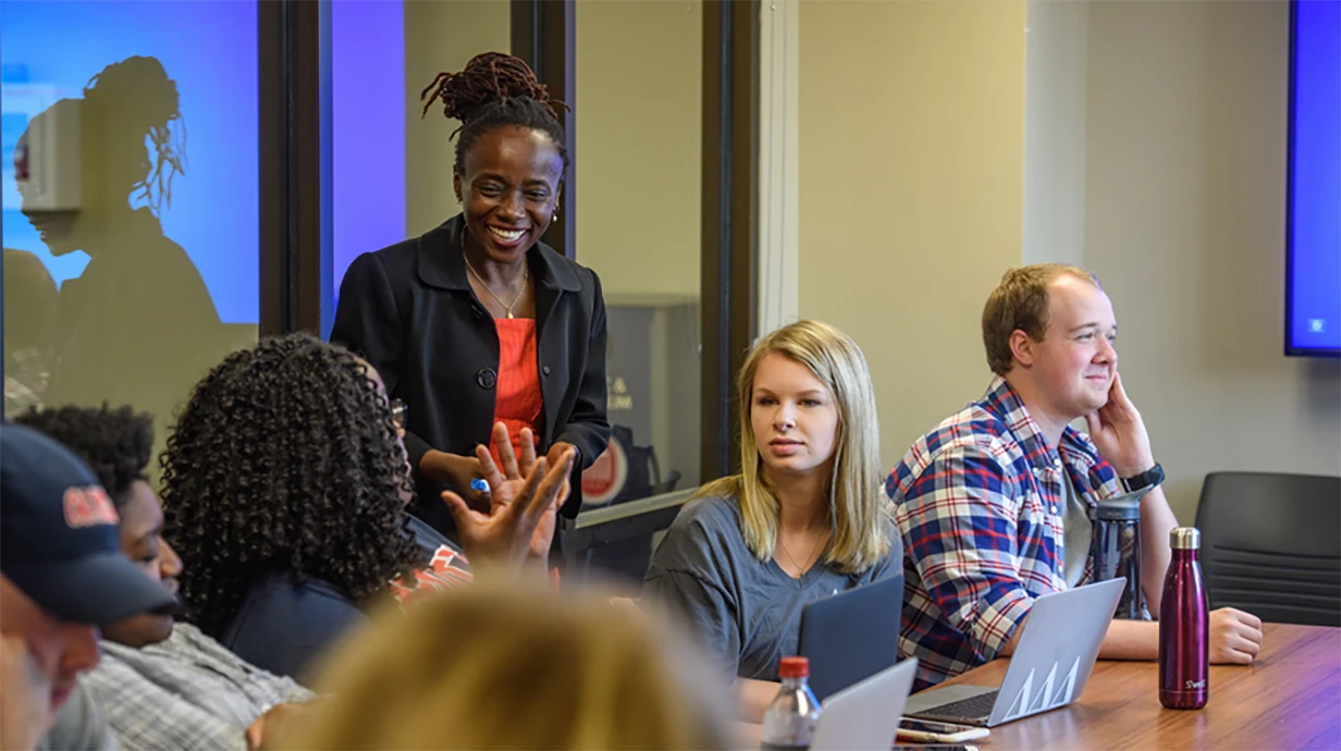 A diverse group in a meeting room. A smiling Black woman with dreadlocks gestures while speaking to attentive colleagues, including a blonde woman and a man in plaid shirt.