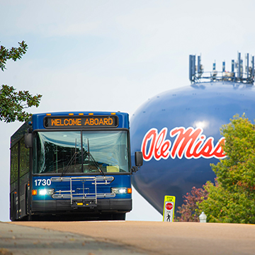 An OUT blue bus drives over a hill with the Ole Miss water tower in the background, and a "Welcome Aboard" message displays across the digital screen on the front of the bus