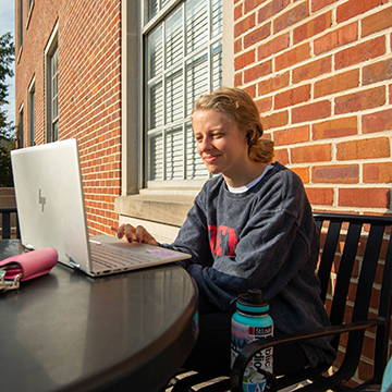 A student looks at her laptop on the front porch of the library while sitting at a table