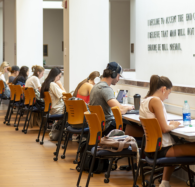 Many students sit at a long desk in the library and study during finals week