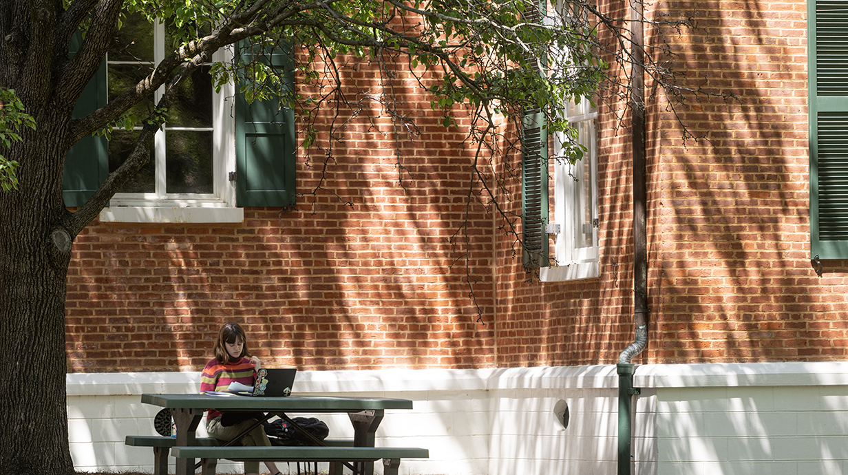 A student sits alone at a picnic table under a tree in the grove with a red brick building behind them