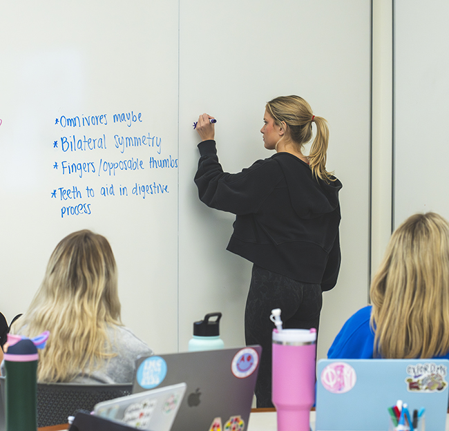 A student stands at a whiteboard and writes down important notes as her classmates watch and take notes.