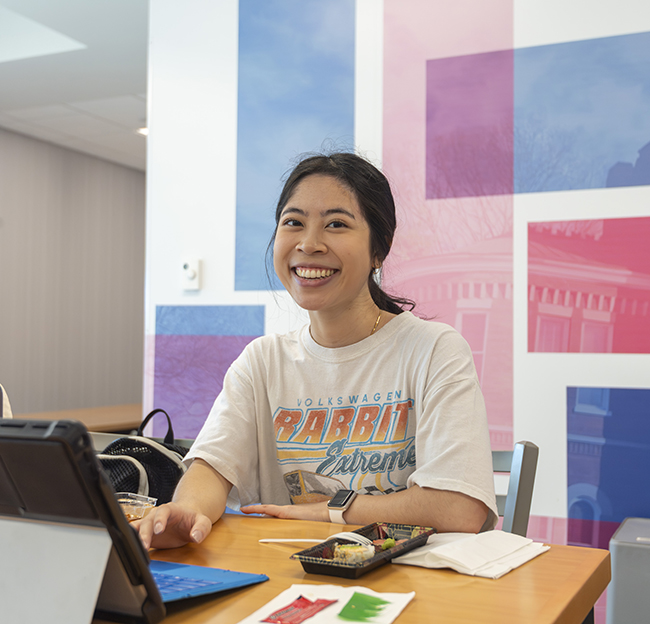 A student smiles and looks at the camera with her laptop and sushi in front of her while studying for finals week.