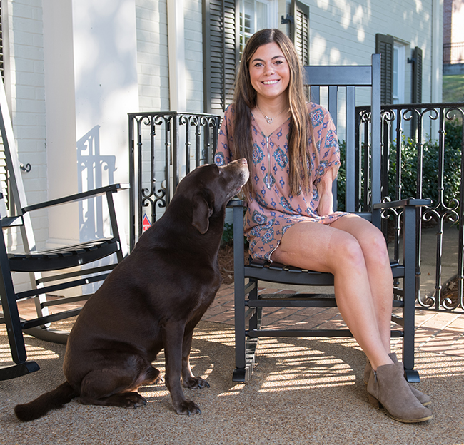A student sits in a rocking chair with her service dog lovingly looking up at her while she smiles at the camera.