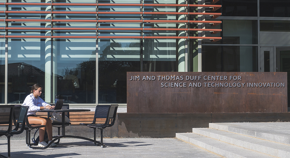 A student with a laptop sits outside a large building.