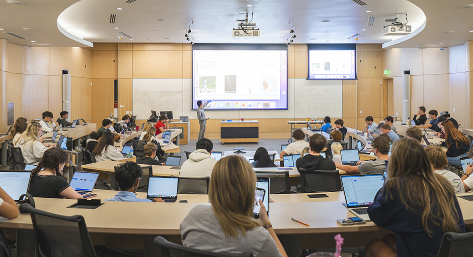 An instructor stands at the front of a large classroom pointing out key elements on a projection screen.