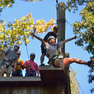 A student zip lining at the challenge course with other students waiting on top of the tower. 