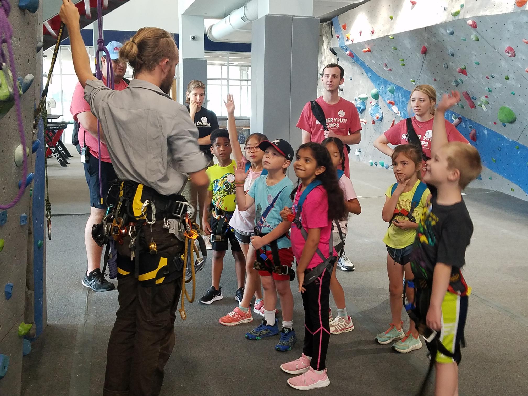 Group of children wearing climbing harnesses are listening to a person instructing them on using a climbing wall. One child has his had raised.