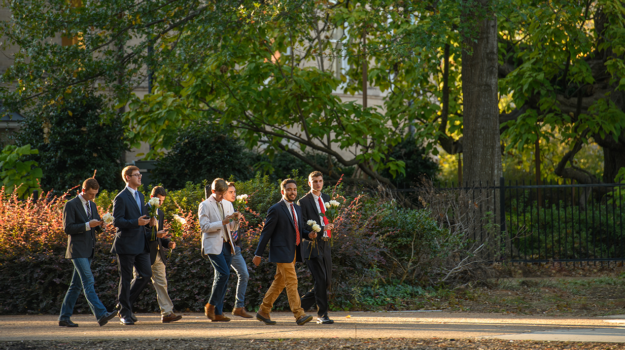 Seven IFC members dressed in jackets and ties carry flowers to a sorority