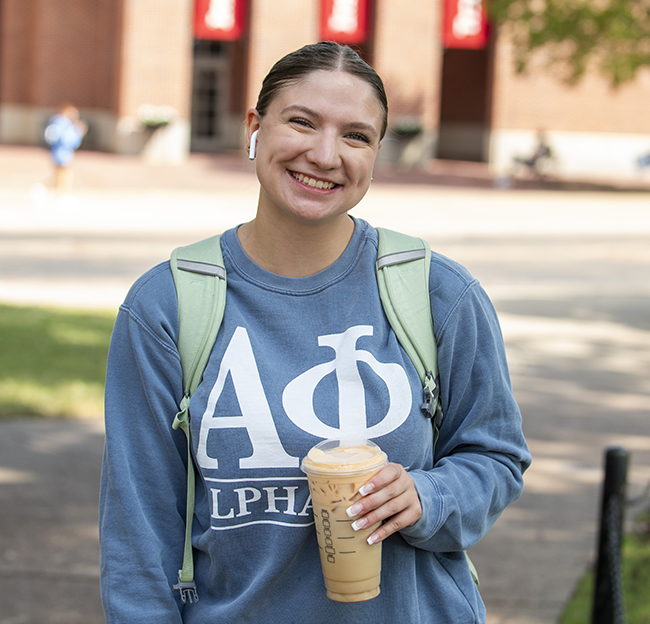 A student wearing an Alpha Phi sweatshirt holds her coffee and smiles