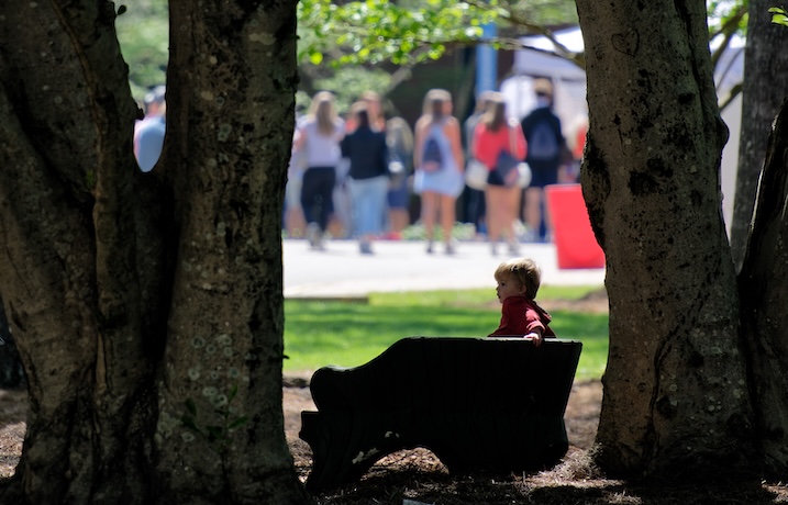 A group of people stand in the background while a child on a bench looks on in the foreground. 