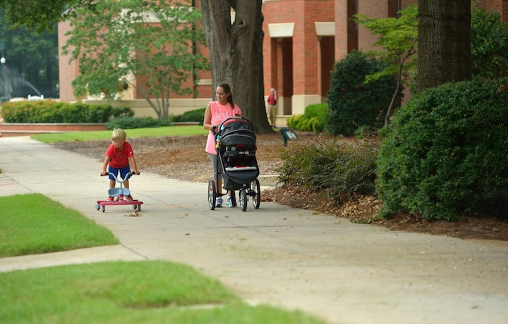 A mom pushes a stroller across campus while watching her child ride a bike.