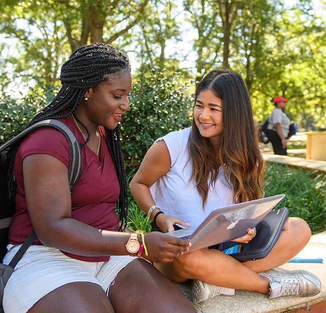 Two students smile as the one on the left passes her laptop to the one on the right as they sit on the brick surrounding the flagpole in the circle