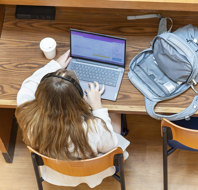 A student sits at a desk with headphones on and looks at her laptop.