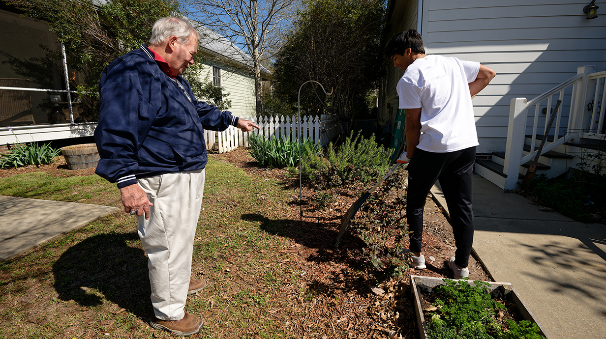 A man wearing a blue windbreaker directs a young man spreading mulch in a landscaped area.