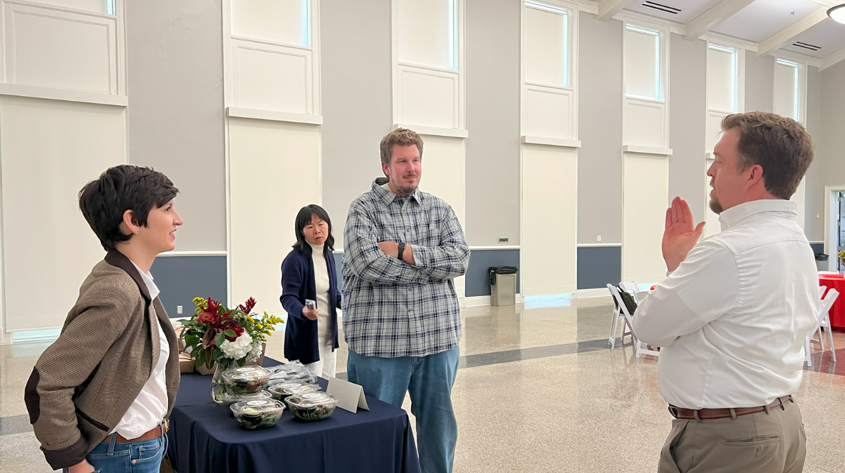 Four people are having a conversation in a bright, spacious room. One person stands by a table with salads and flowers, while another gestures as they speak. Two others watch the interaction nearby. The room has high ceilings and large windows, giving it an open feel.