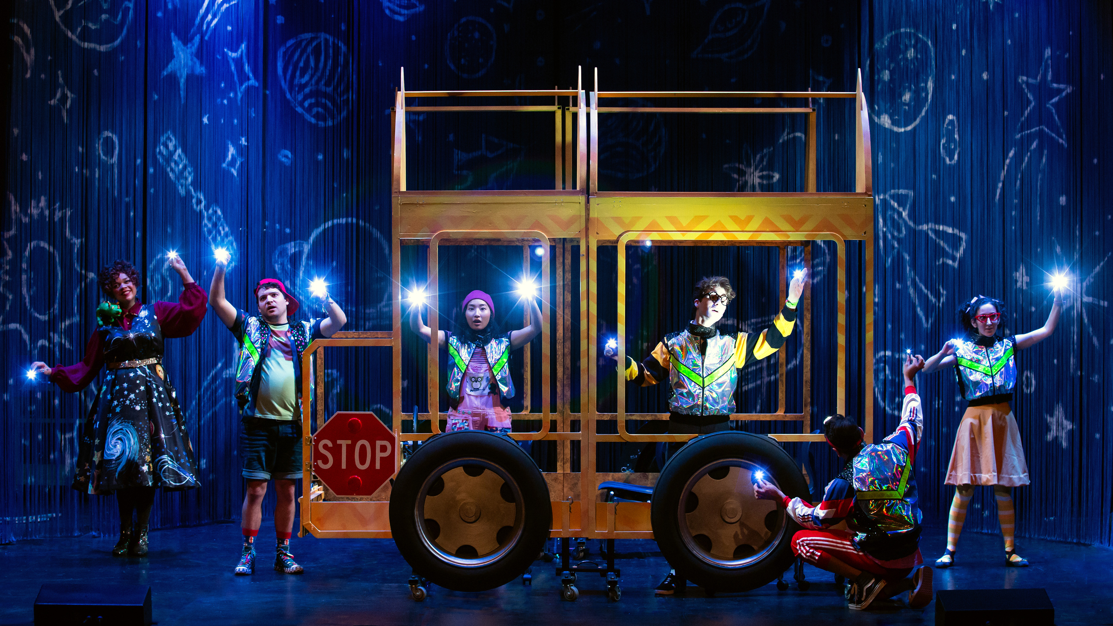 Six actors perform on a theater stage arranged around the skeletal yellow frame of a school bus with large tires and a stop sign. The backdrop is dark blue, decorated with chalk-like drawings of planets, stars, and rockets. Five performers stand spread across the stage wearing colorful, reflective clothing and shining small blue lights directly toward the audience, while one performer kneels near a tire in the foreground.