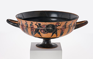 Ancient Greek black-figure kylix with two horizontal handles, decorated around the exterior with silhouetted figures of soldiers and athletes engaged in combat and boxing, and a central interior scene of a running youth, shown on a pedestal against a white background.