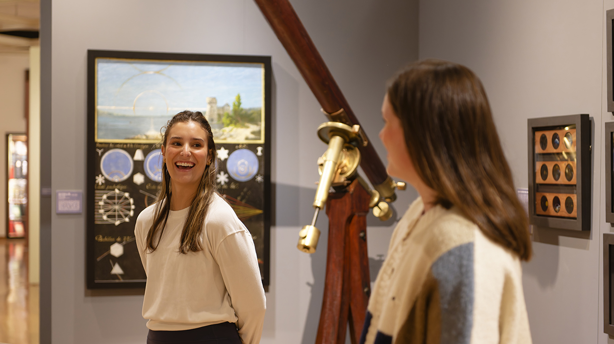 Two visitors talk and smile inside a museum gallery, standing near a large antique telescope and scientific instruments, with framed exhibits displayed on the walls behind them.