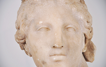 Close-up of a classical marble sculpture head, showing a serene female face with softly carved features, wavy hair pulled back, and weathered stone surface against a neutral background.