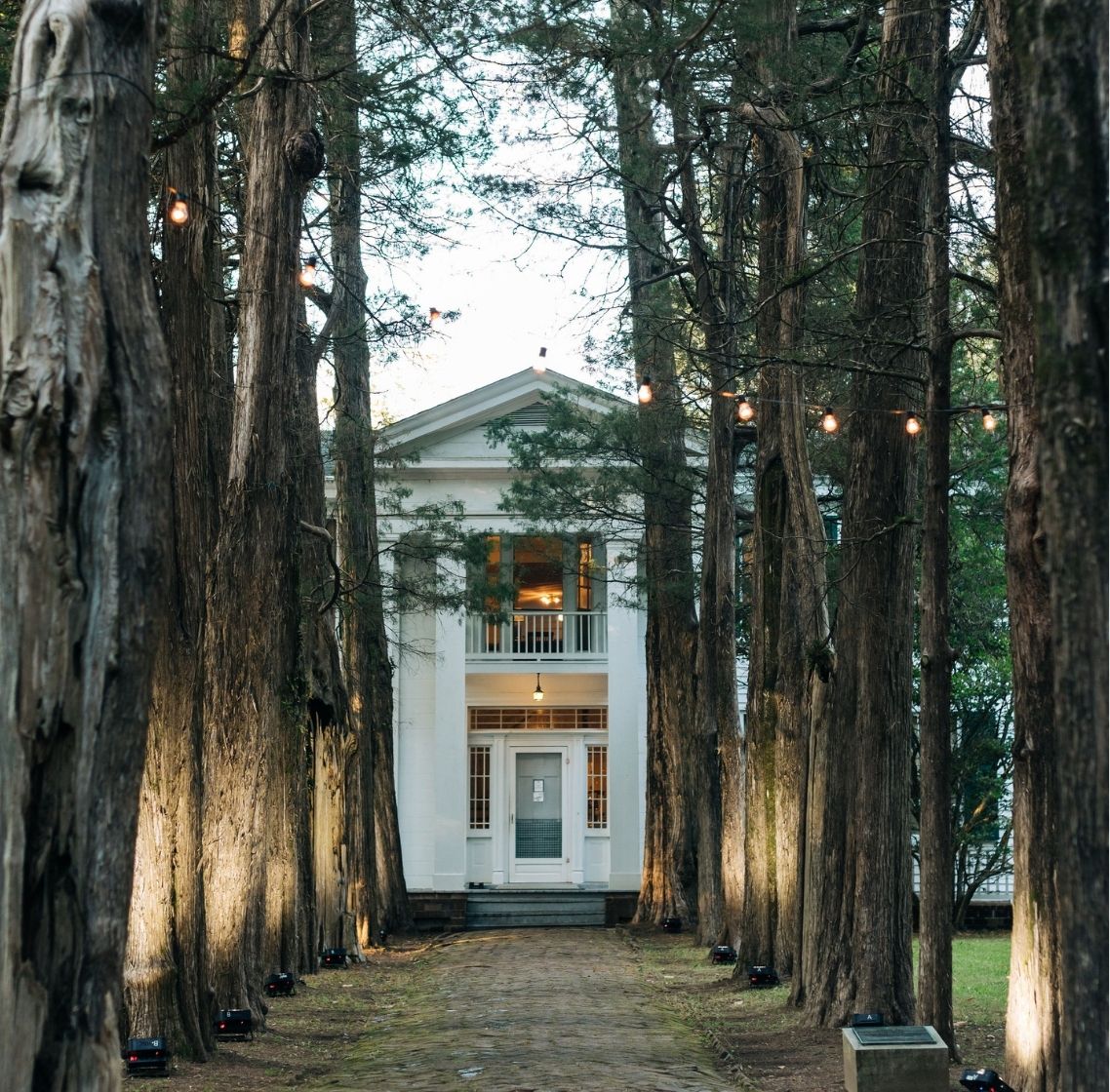 Rowan Oak’s white two-story facade partially framed by tall trees with string lights hanging among the branches.
