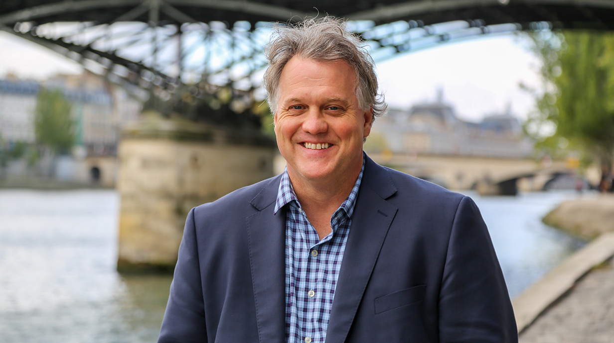 A man wearing a blue jacket stands in front of a bridge.