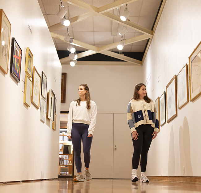Two women walk through a museum gallery, viewing framed artworks displayed along white walls.