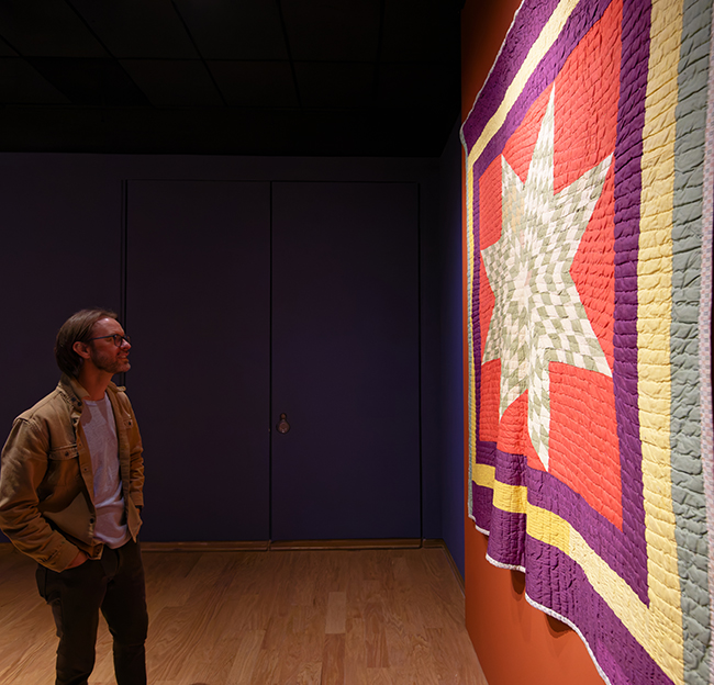 Man viewing a large, colorful star-pattern quilt in a dimly lit museum gallery.